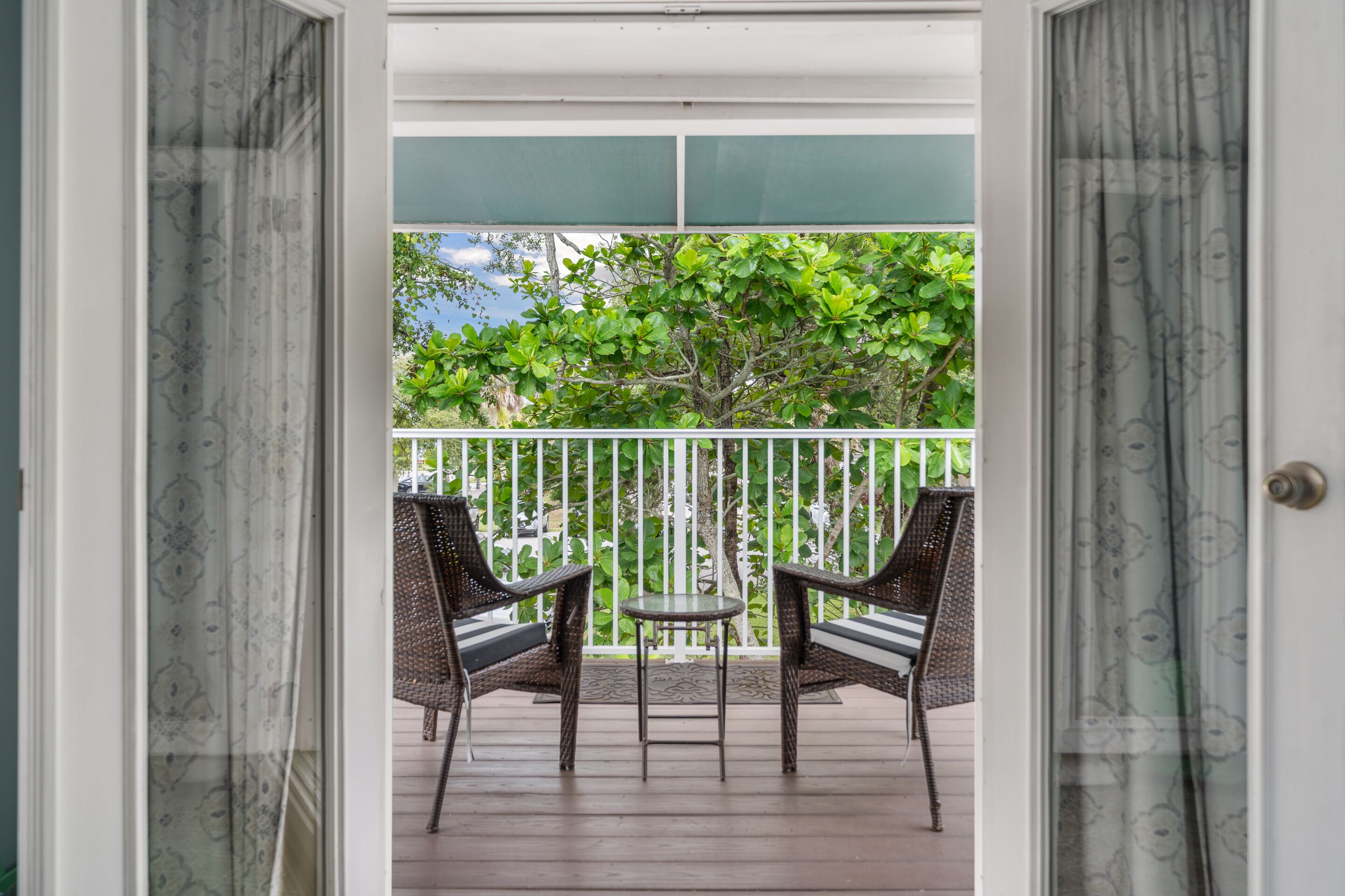 A balcony with two wicker chairs and a small table, surrounded by a white railing and a view of greenery.