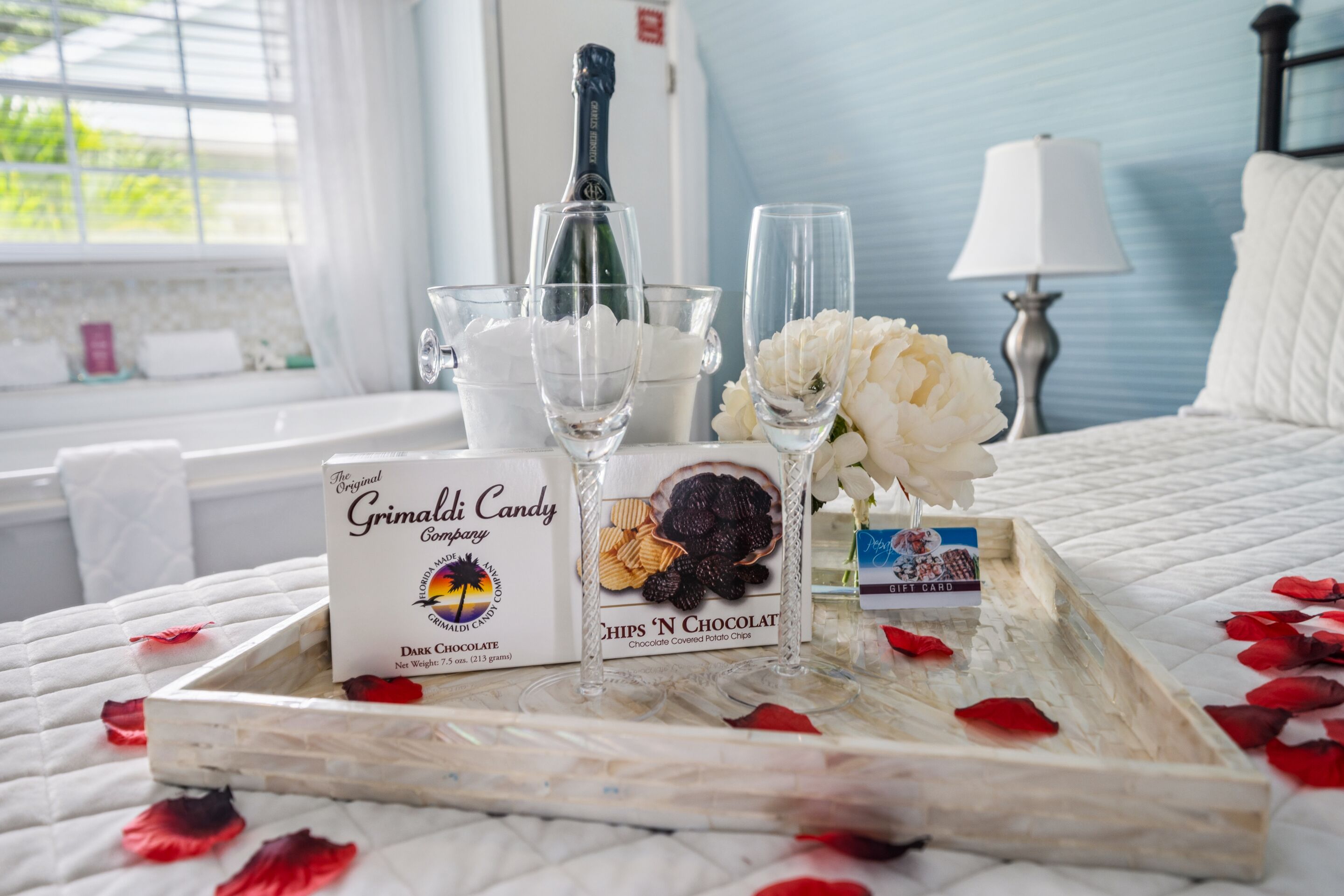 A tray with Grimaldi Candy, a bottle of champagne, and a gift card on a bed with white bedding and rose petals.