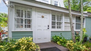 Une maison verte avec une porte et des fenêtres blanches, entourée de plantes tropicales et agrémentée d’un paillasson.