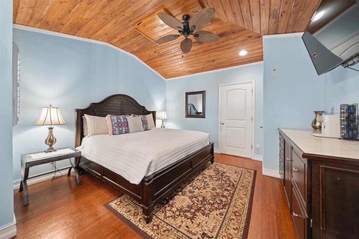 Um quarto com teto de madeira, uma cama com colcha branca, duas luminárias de cabeceira, um tapete estampado e um armário de madeira.