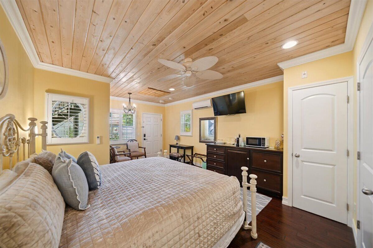 A bedroom with a wooden ceiling, a bed with a quilted bedspread, a dresser, a television mounted on the wall, and a door.