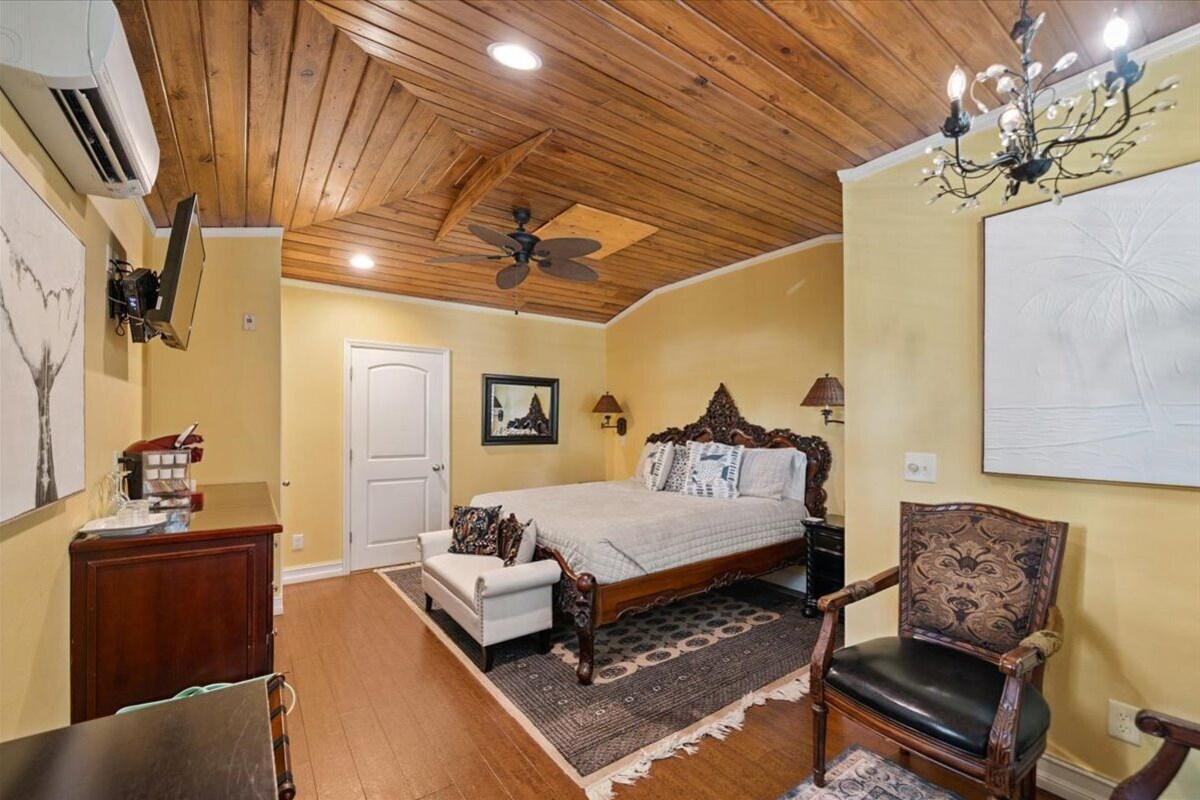 A bedroom with a wooden ceiling, a bed, a chair, a desk, and a chandelier.