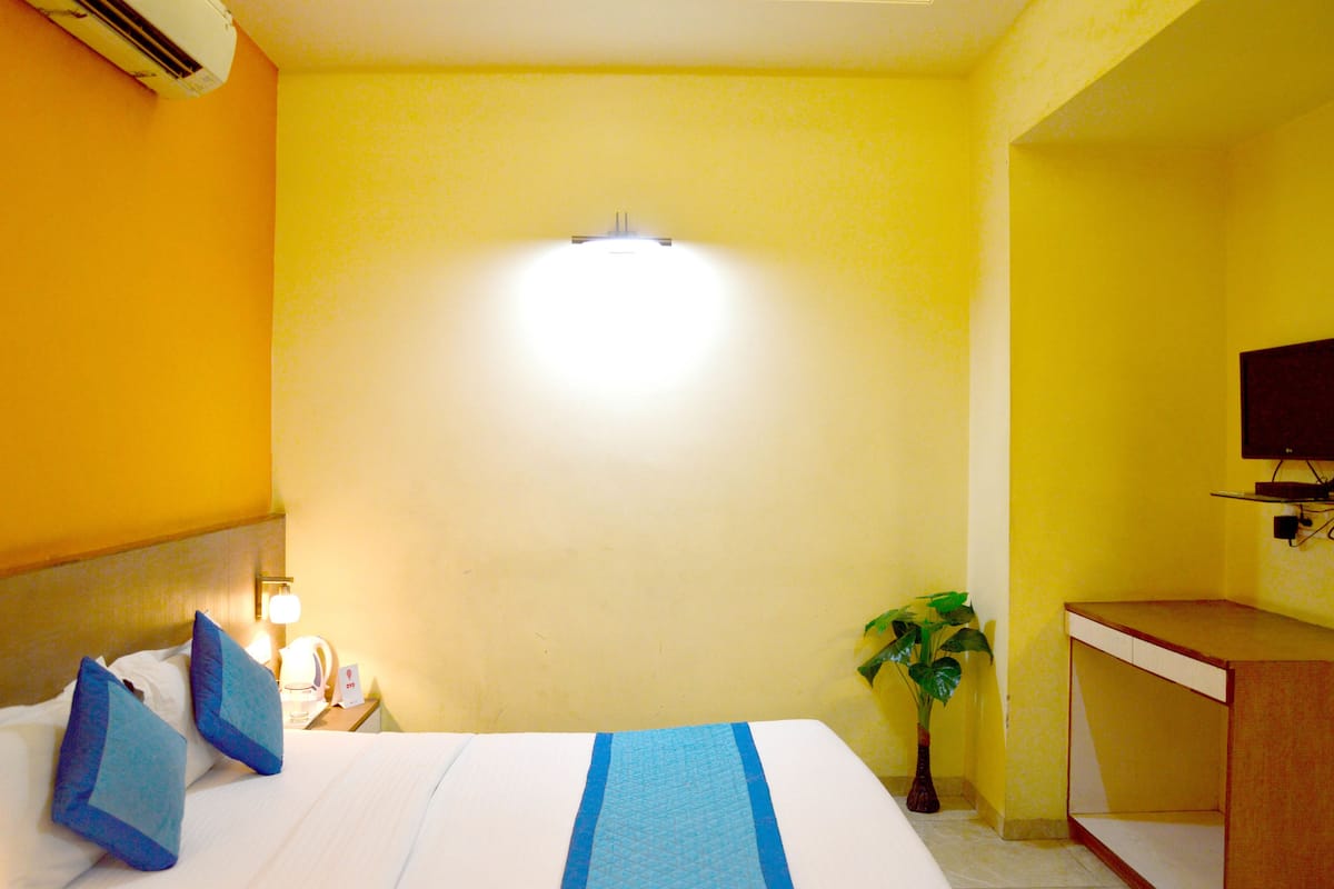 A hotel room with a bed, a bedside table with a lamp, a small plant, and a desk with a chair.
