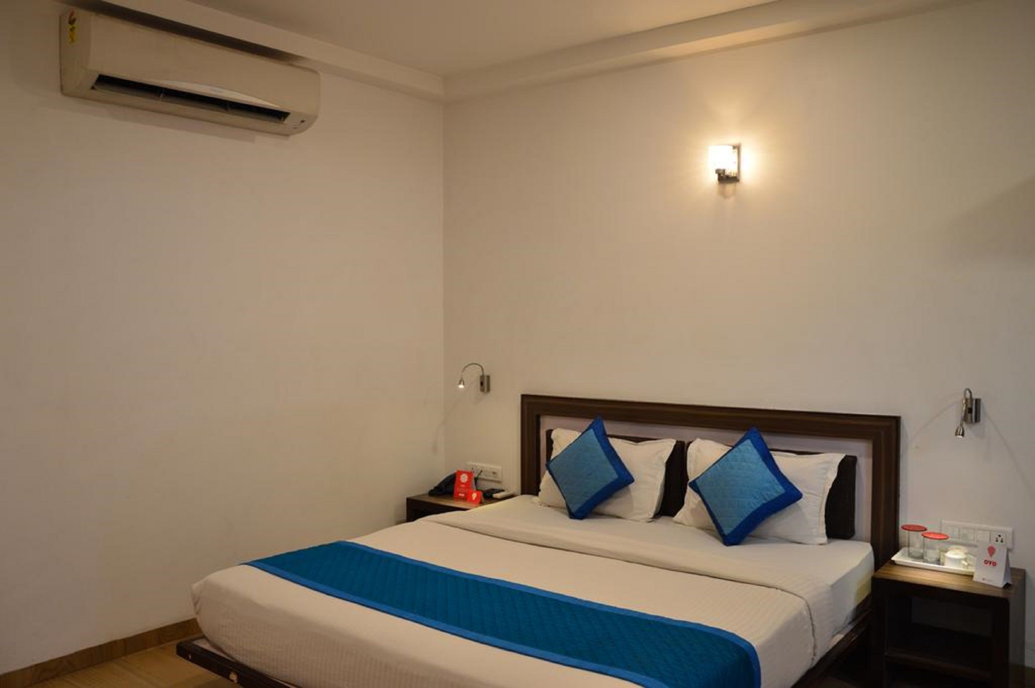 Una habitación de hotel con una cama, una mesita de noche, un equipo de aire acondicionado y una lámpara de pared.
