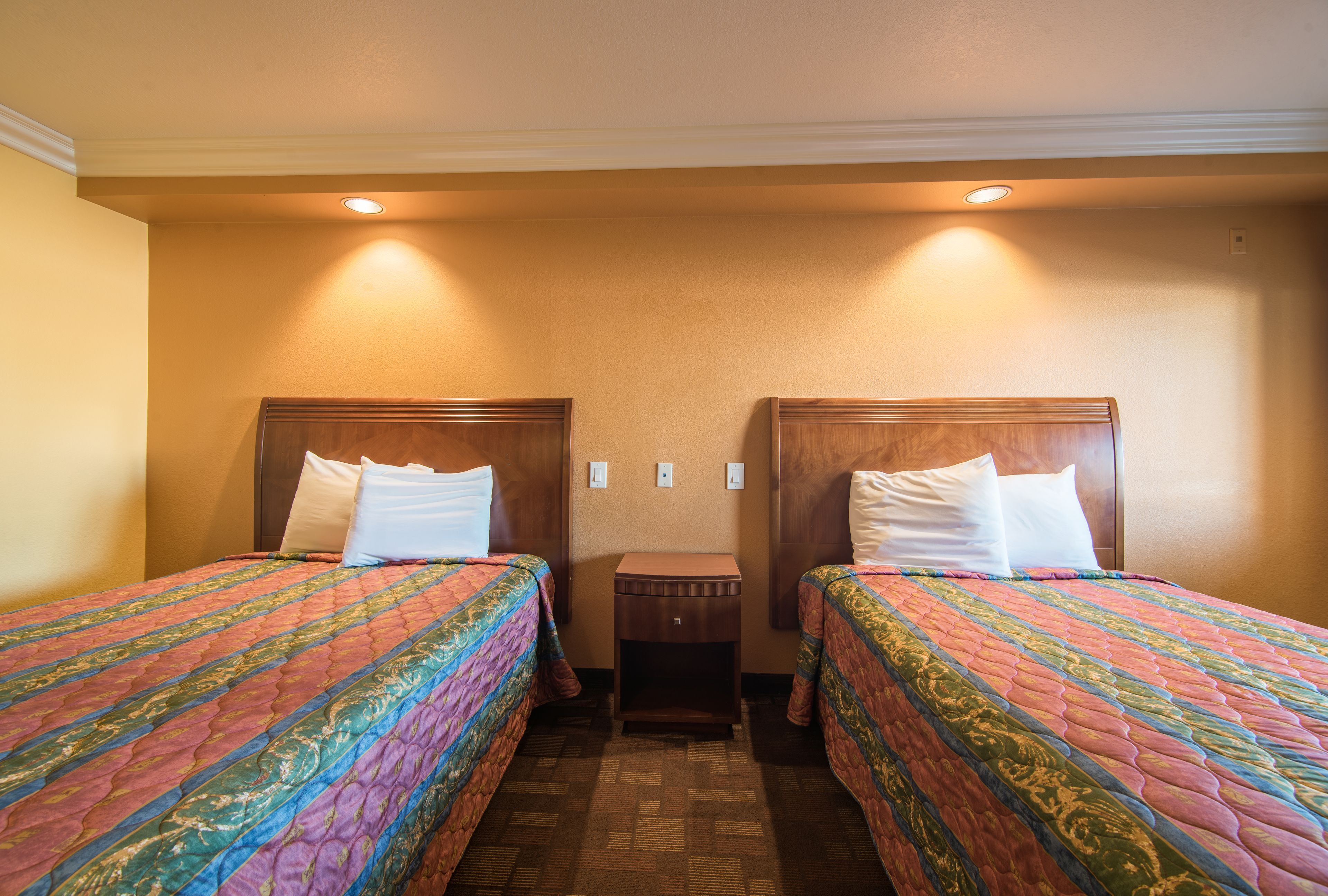 A hotel room with two single beds, each with a wooden headboard and a colorful quilt. There is a small wooden nightstand between the beds.