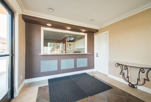 A hotel entrance with a reception desk, a black doormat, a glass panel wall, and a white door.