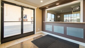 A reception area with a glass partition, a reception desk, a set of double doors, and a doormat. - Industry Inn & Suites (La Puente)