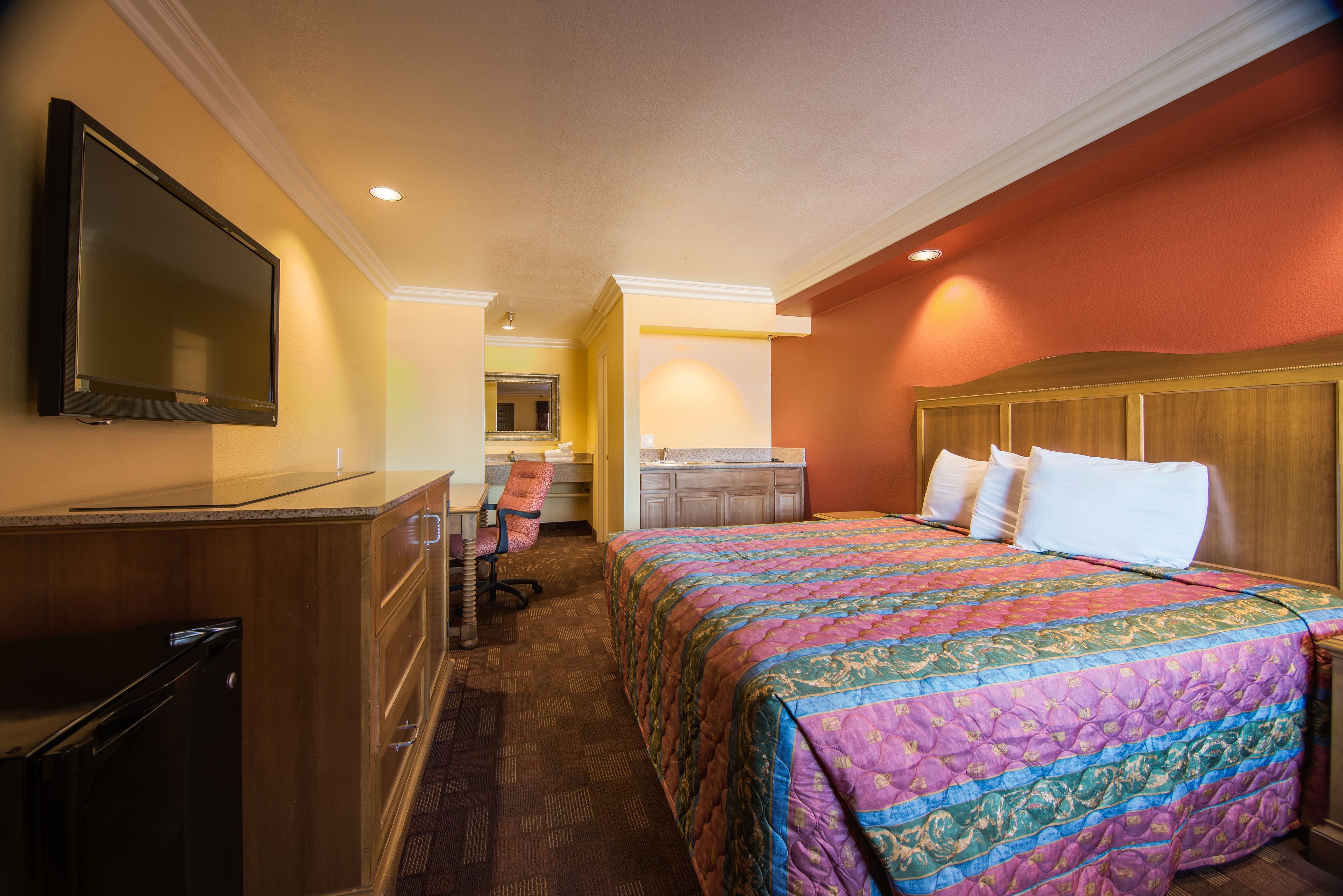 A hotel room with a large bed, a TV mounted on the wall, a desk with a chair, and a kitchenette in the background.