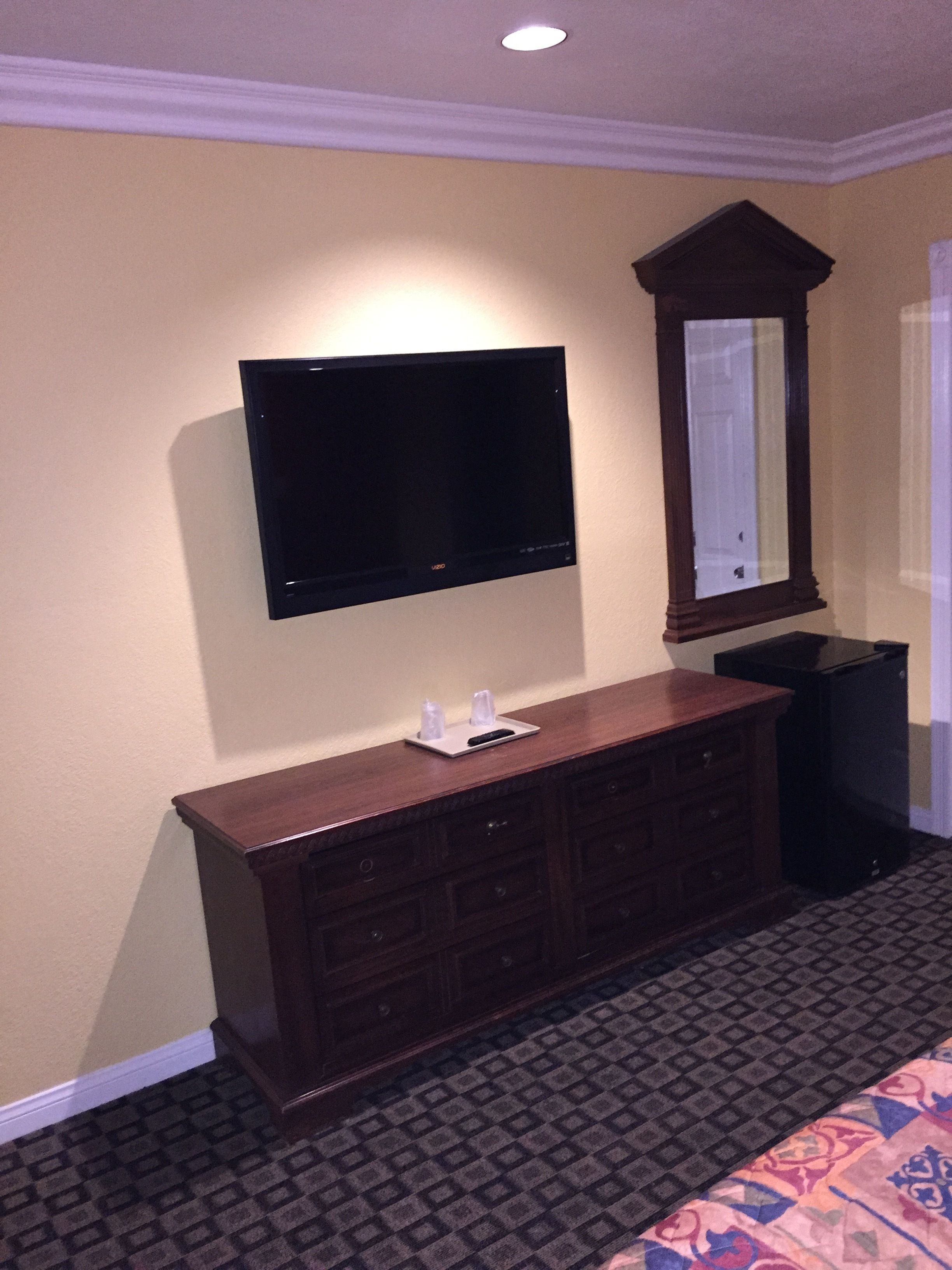 A hotel room with a flat-screen TV mounted on the wall, a wooden dresser with a mirror above it, and a patterned carpet.
