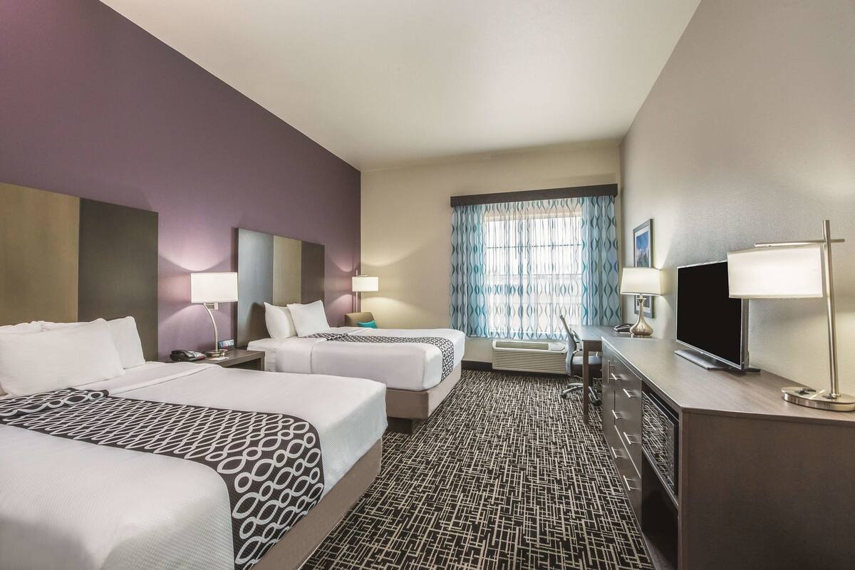 A hotel room with two beds, a desk, a TV, and a window with curtains.