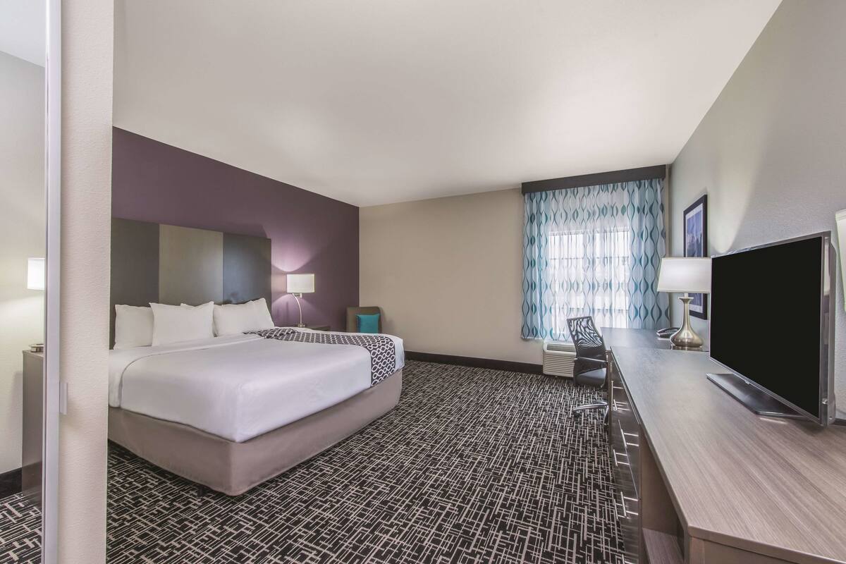 A hotel room with a large bed, a desk with a flat-screen TV, a chair, a desk lamp, and a window with patterned curtains.