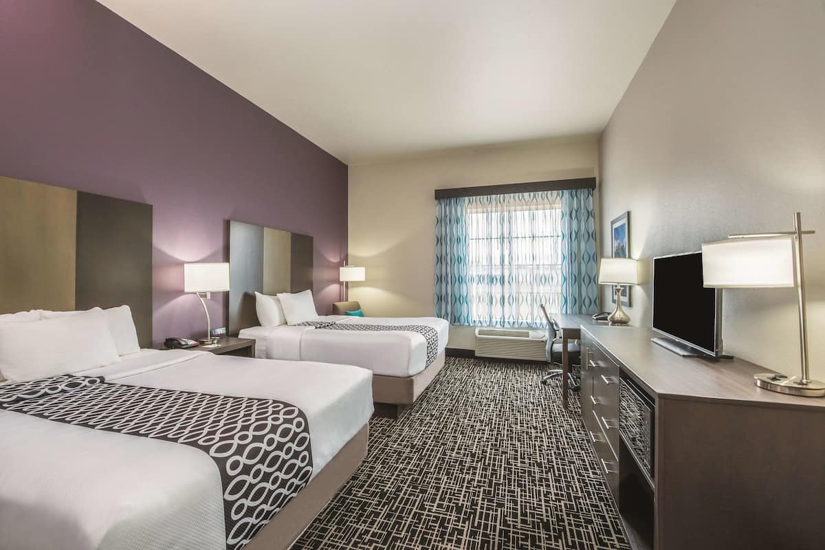 A hotel room with two beds, a desk with a TV, and a window with patterned curtains.