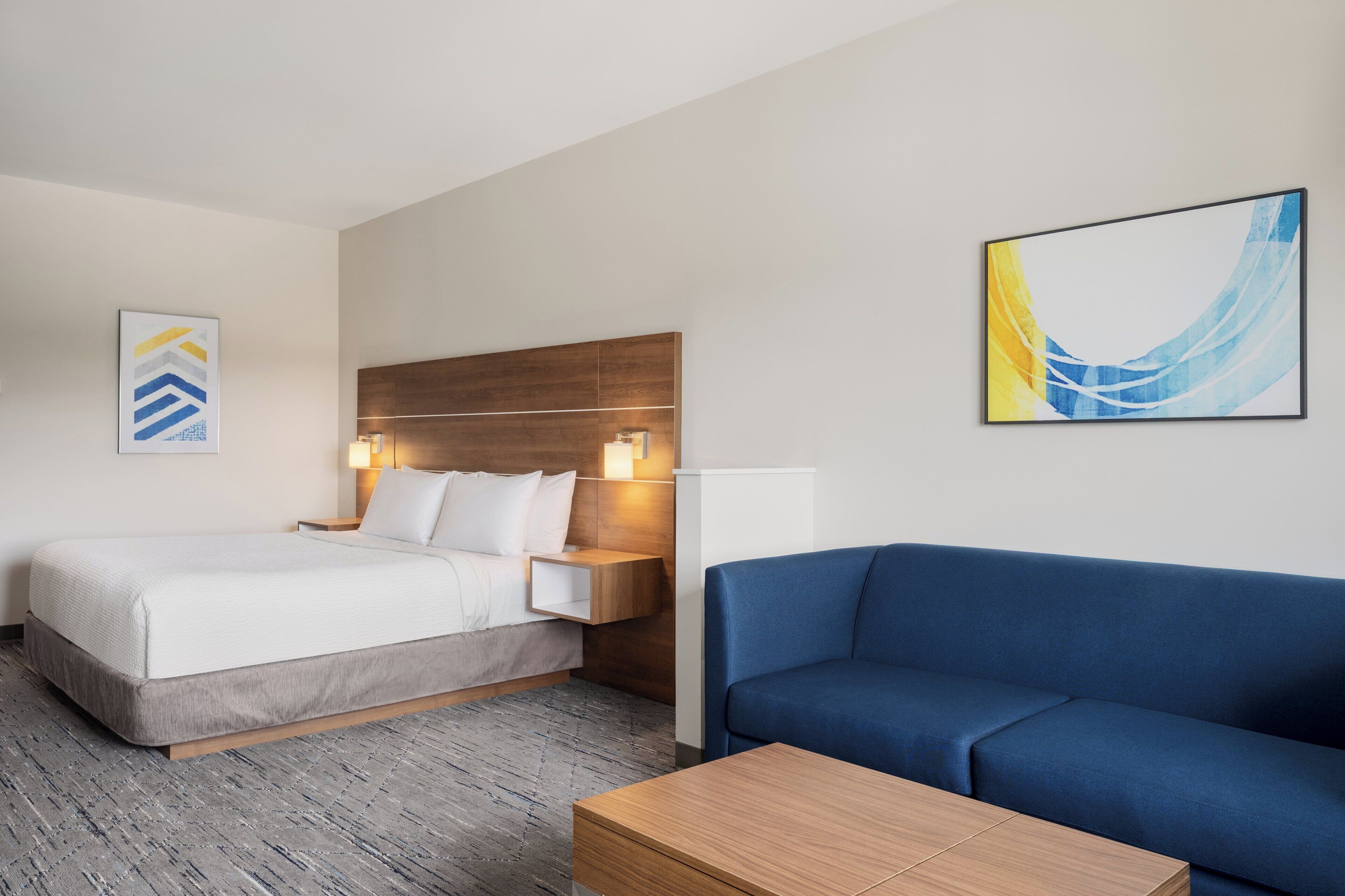 A hotel room with a bed, a blue sofa, a wooden coffee table, a nightstand with a lamp, and two wall-mounted artworks.