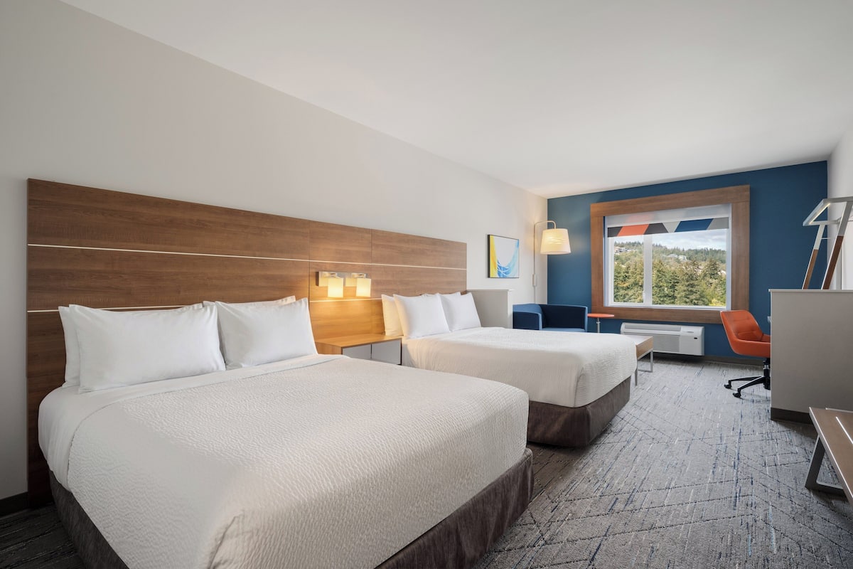 A hotel room with two beds, a desk, a chair, a window with a view, and a lamp.