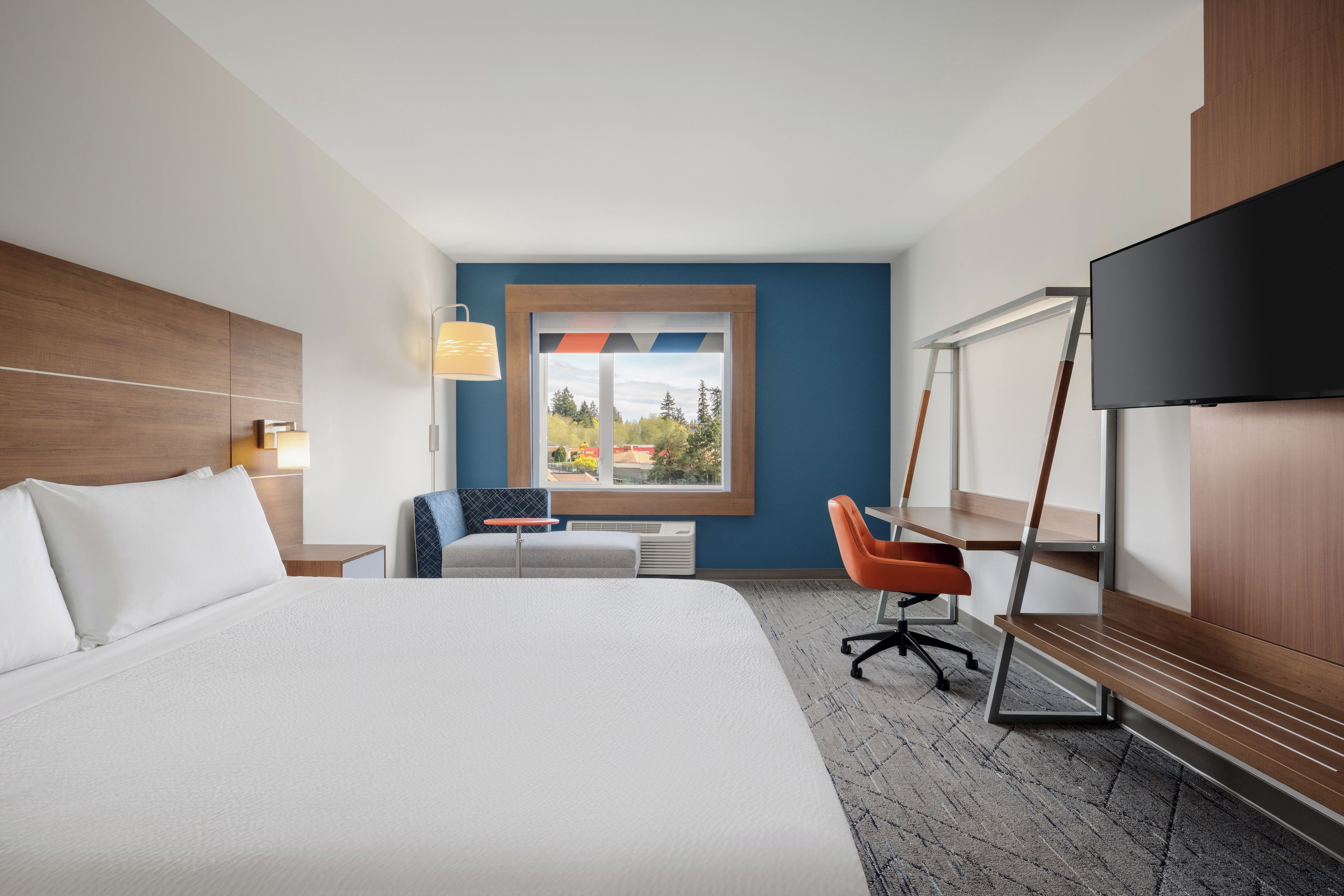 Habitación de hotel con una cama grande, un escritorio, una silla, un televisor y una ventana con vistas a árboles y edificios.