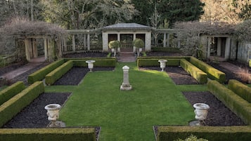 Un jardín formal con césped central, setos simétricos y una estructura de edificio de estilo clásico.