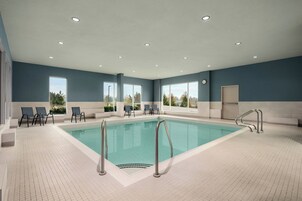 Una piscina interior con agua cristalina y azul, rodeada de sillas de descanso y un suelo de baldosas.