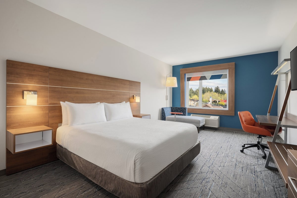 A hotel room with a large bed, a desk with an orange chair, a window with a view, and a wooden headboard.