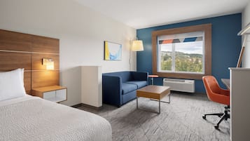 Quarto de hotel com cama, sofá, escrivaninha, cadeira, abajur, radiador e janela com vista.