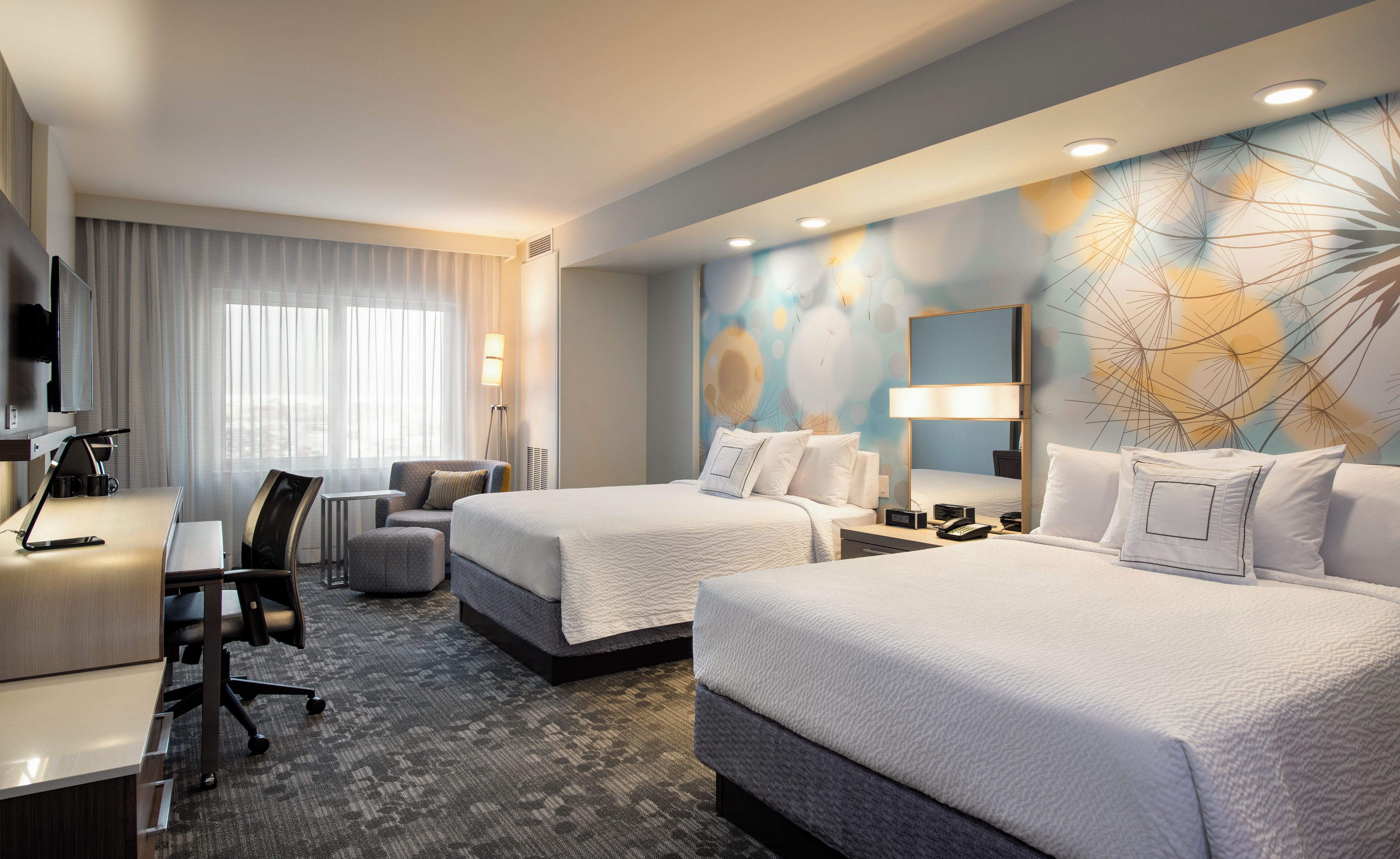 A hotel room with two beds, a desk, a chair, a TV, and a large window with curtains.