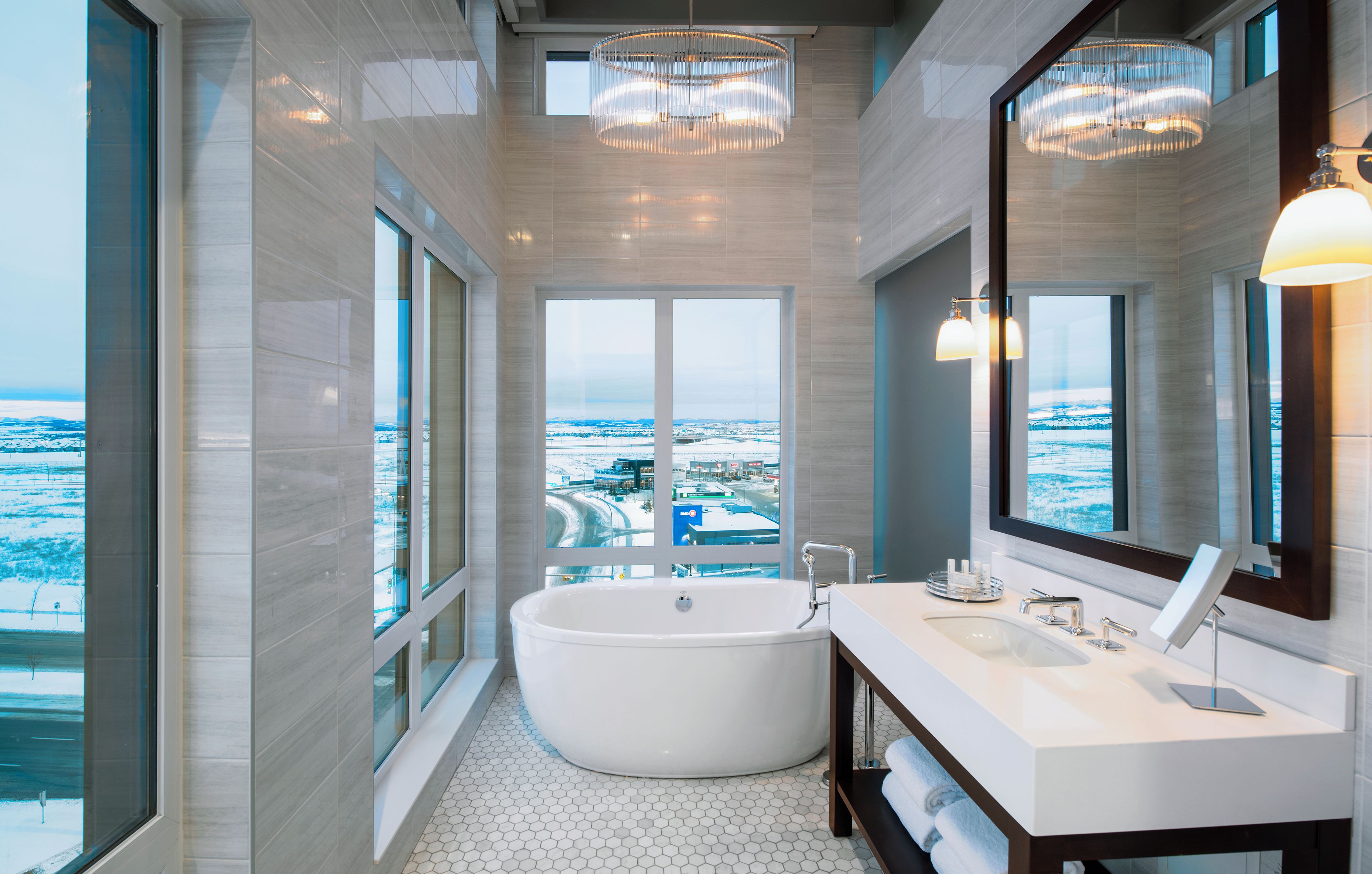 Un baño moderno con una bañera independiente, un espejo grande y vistas a un paisaje nevado a través de las ventanas.