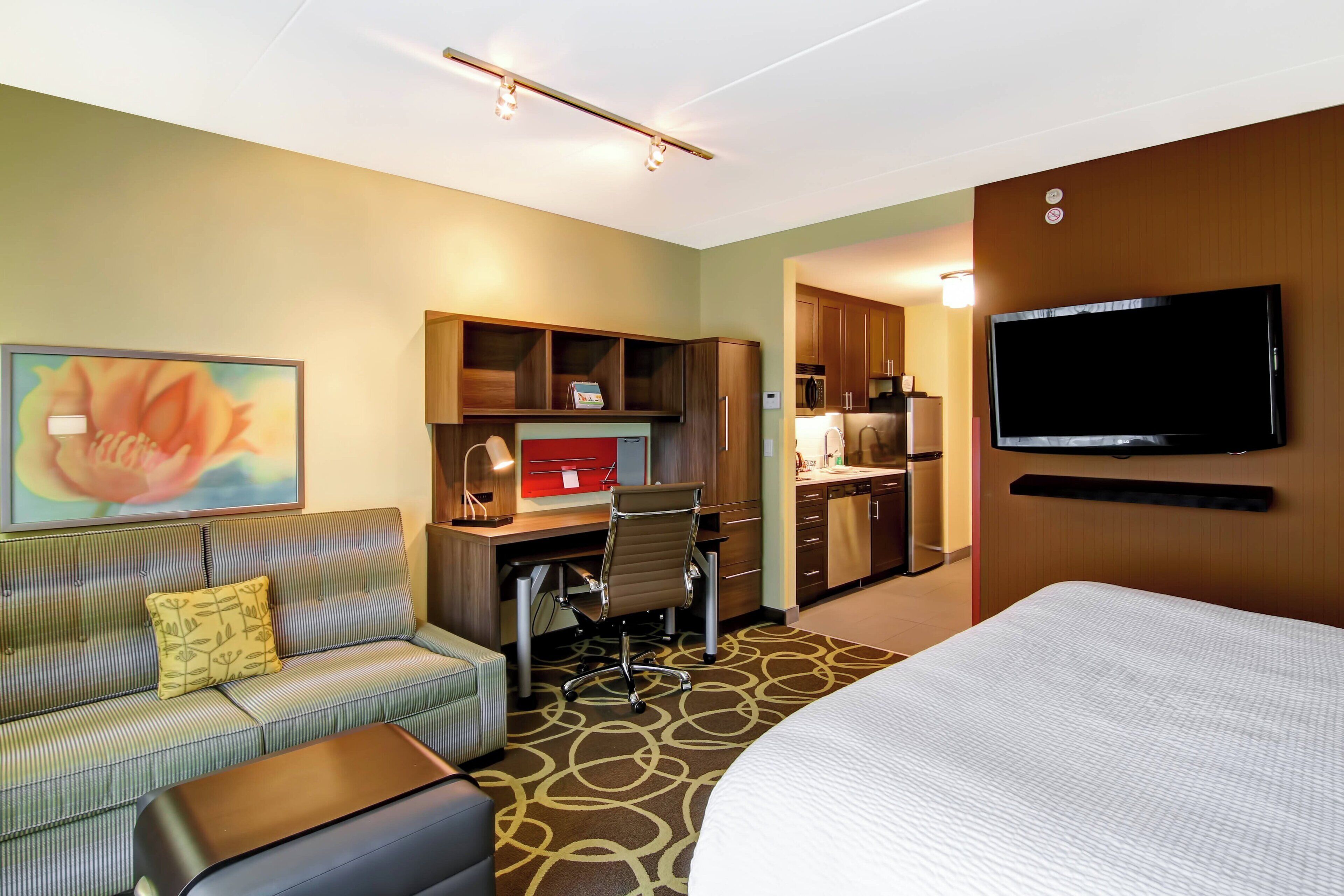 A hotel room with a bed, a sofa, a desk with a computer, a television, and a painting on the wall.