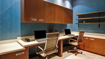 A modern office with three desks, each with a laptop and a chair. There are wooden cabinets above the desks and a shelf on the wall.