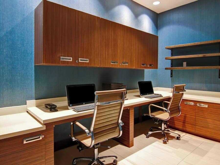 A modern office with three desks, each with a laptop and a chair. There are wooden cabinets above the desks and a shelf on the wall.