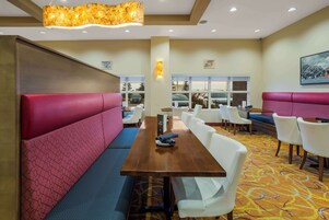 A dining area with a long table, red and blue cushioned seating, white chairs, and a patterned carpet.