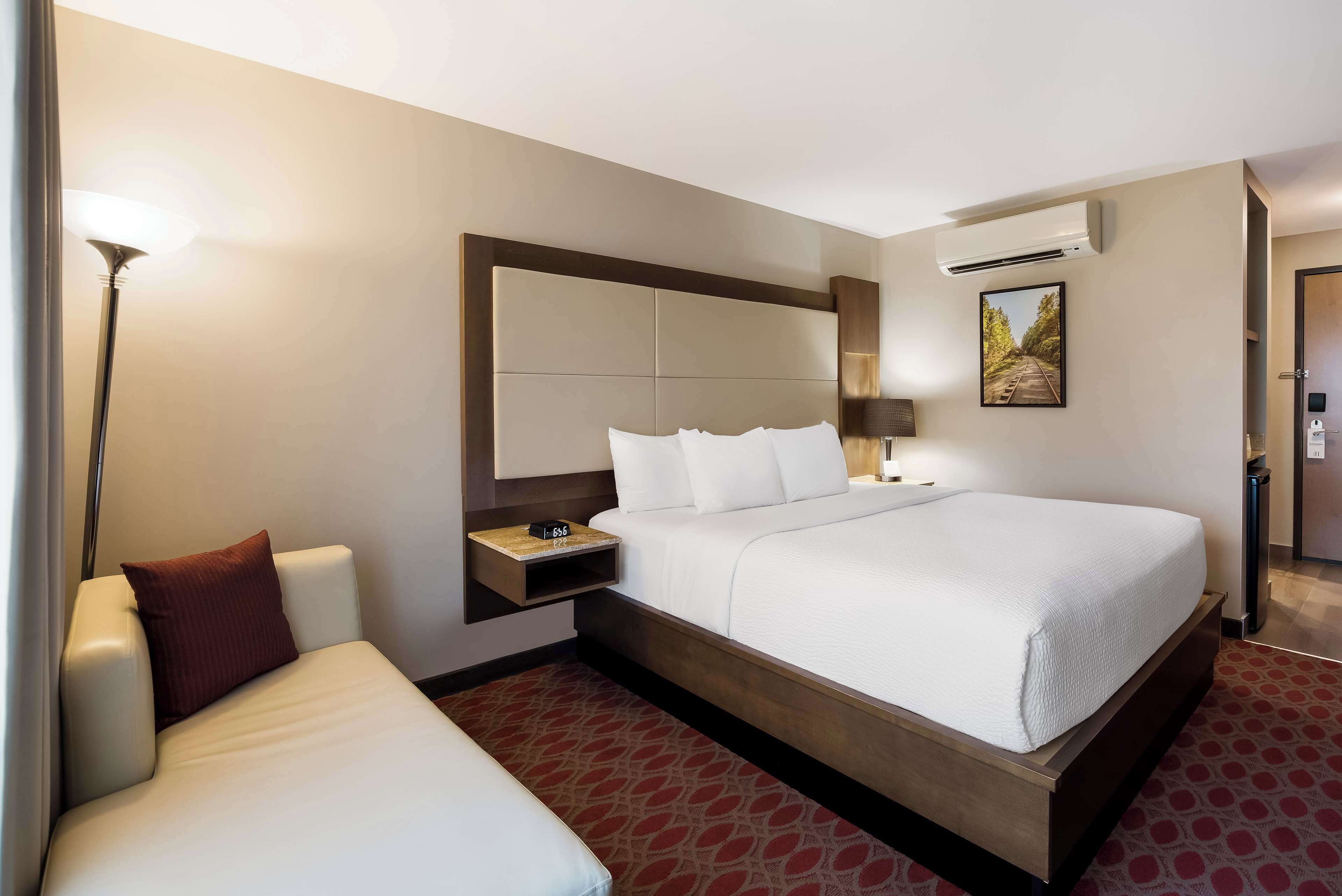 A hotel room with a large bed, a bedside table, a lamp, and an air conditioning unit.