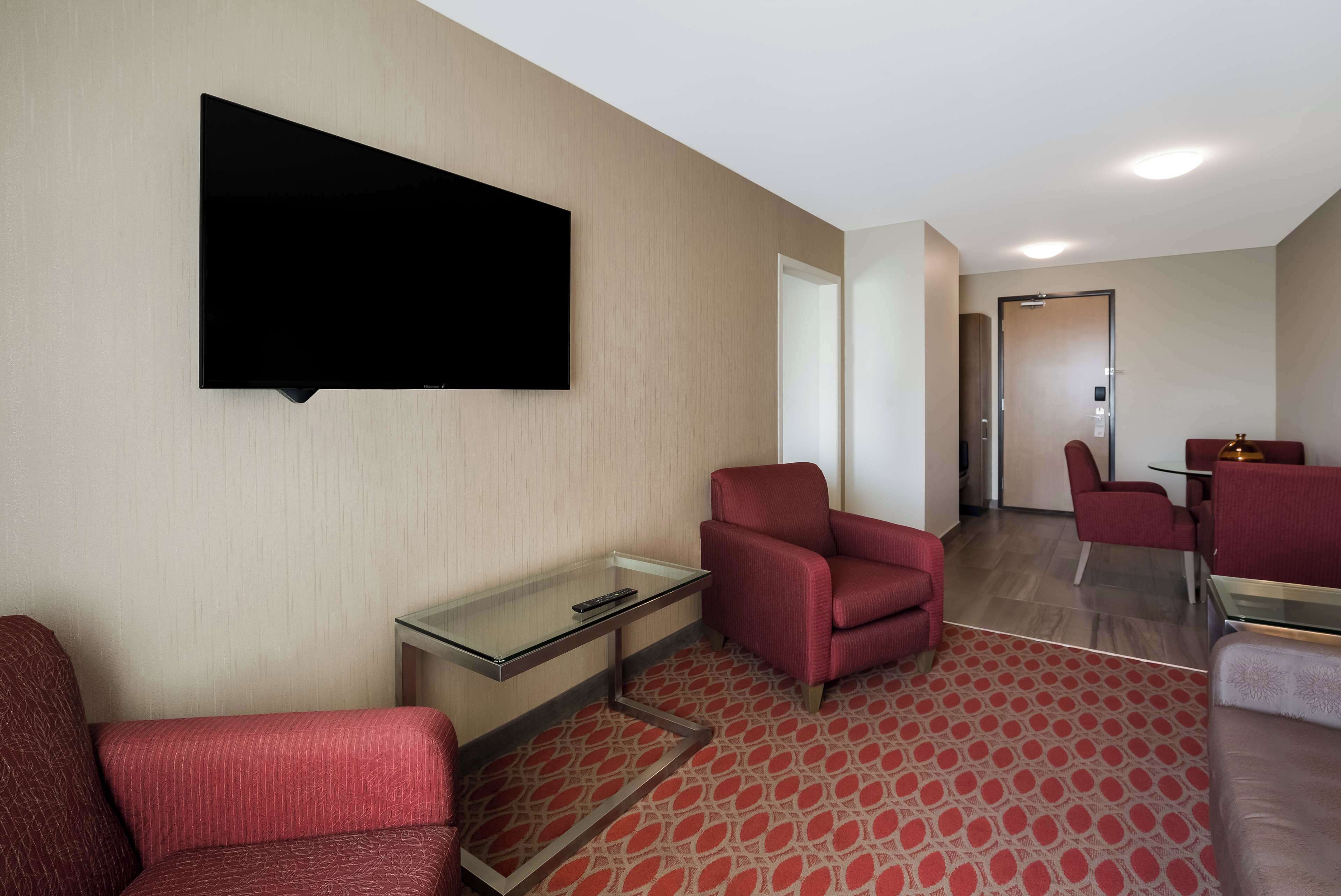 A hotel room with a flat-screen TV mounted on the wall, a glass coffee table, red upholstered chairs, and a patterned carpet.