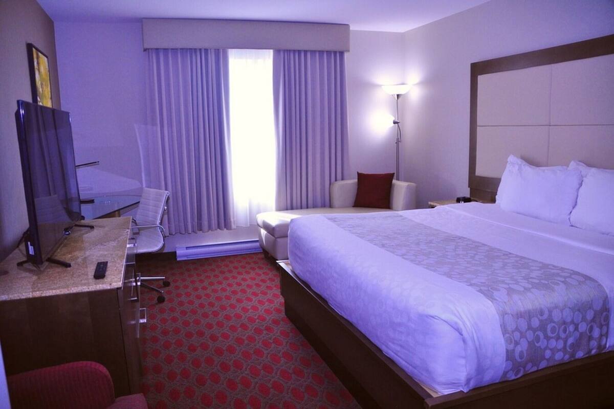 Habitación de hotel con una cama grande, un escritorio con computadora, una silla, una lámpara y un ventanal con cortinas.