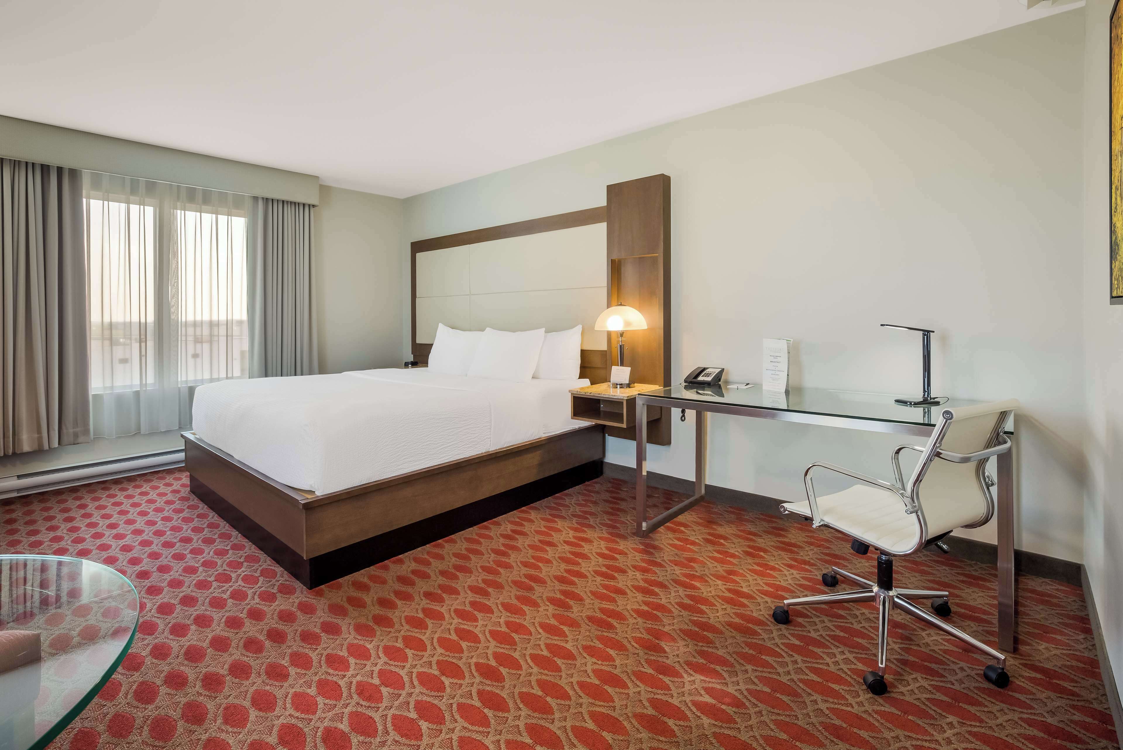 A hotel room with a large bed, a desk with a chair, a bedside table, and a window with curtains.