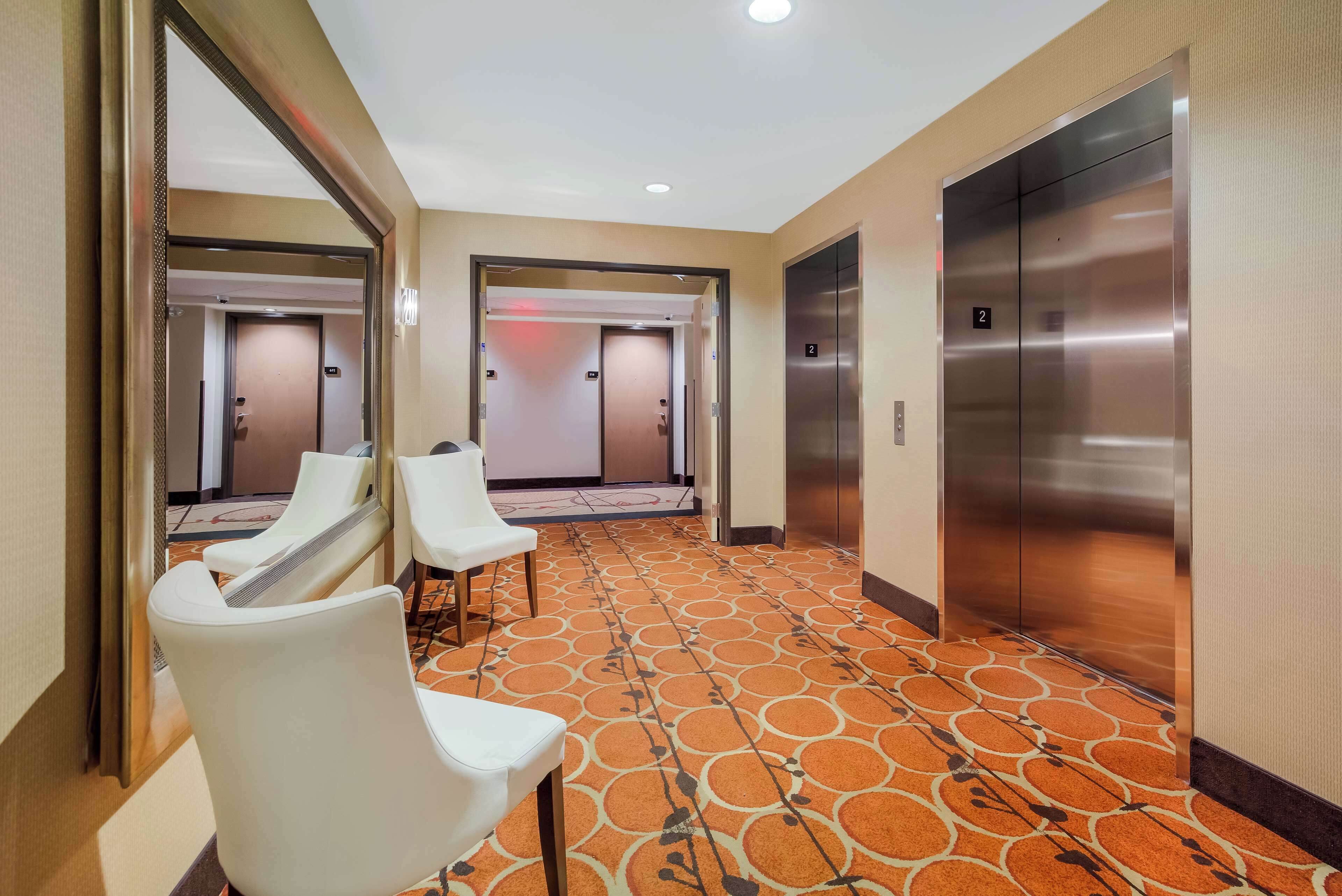 Un pasillo de hotel con dos ascensores, piso alfombrado y sillas blancas.