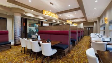 Un espace de restauration d'hôtel avec des banquettes, des chaises blanches et un tapis à motifs.