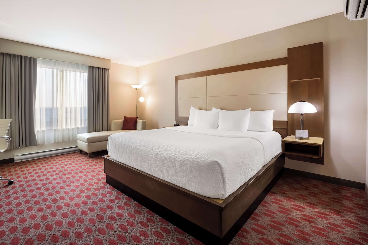 Habitación de hotel con una cama grande, una mesita de noche con lámpara, una silla y una ventana con cortinas.