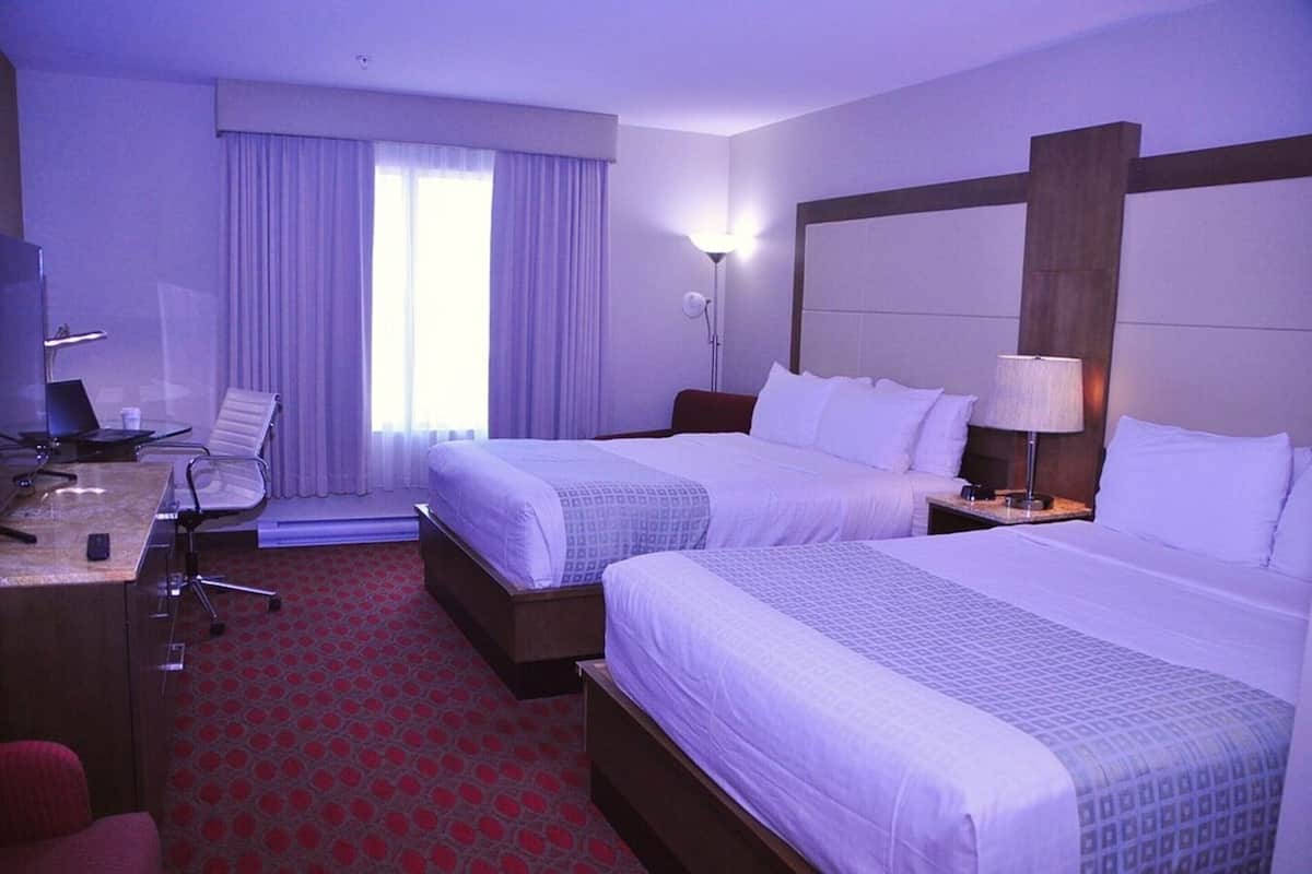 Habitación de hotel con dos camas, un escritorio con silla, una lámpara y una ventana con cortinas.