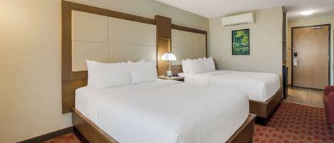 A hotel room with two beds, a wooden headboard, a red patterned carpet, a nightstand with a lamp, and an air conditioning unit on the wall.