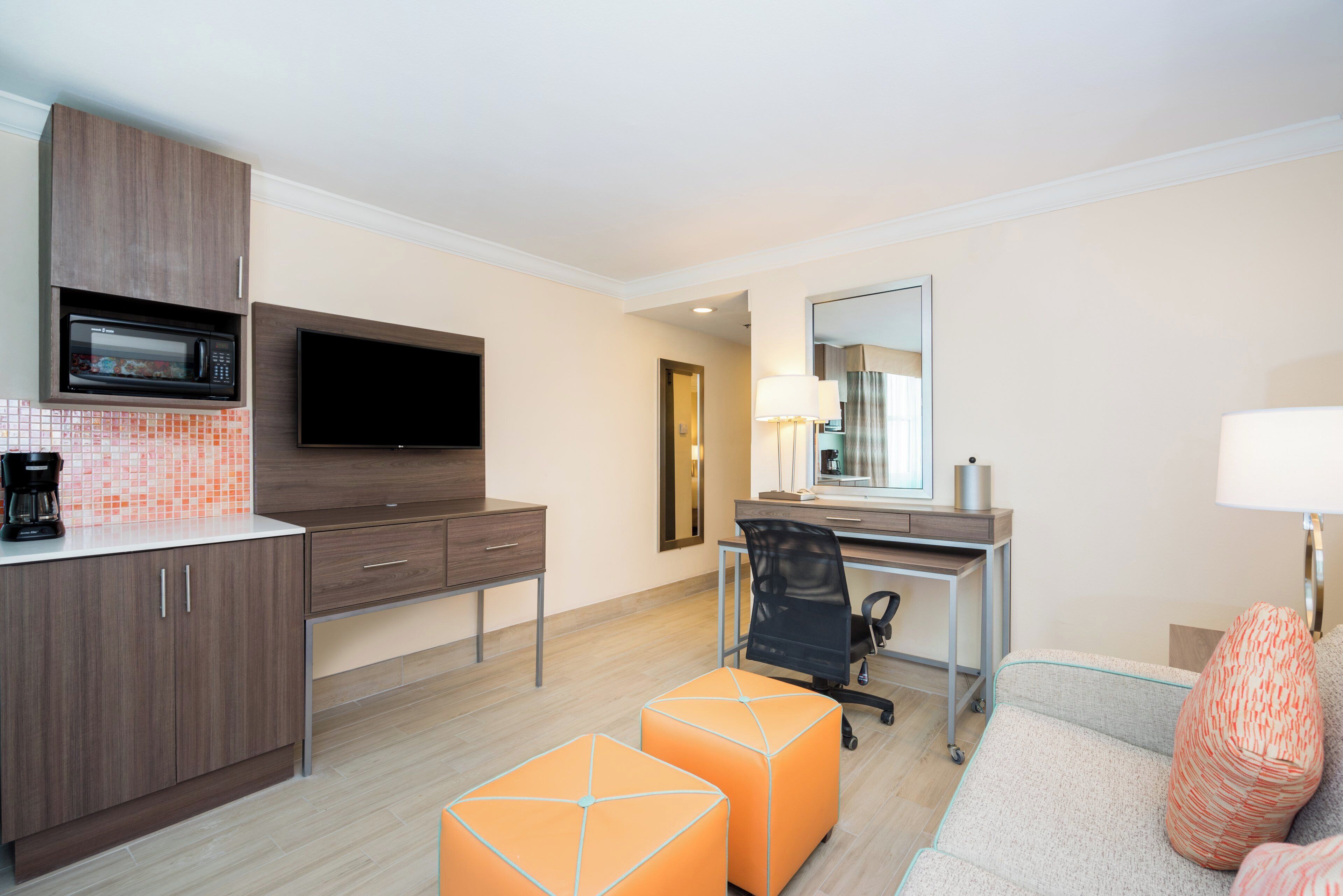 A modern hotel room with a kitchenette, a flat-screen TV, a desk with a chair, and a large mirror.