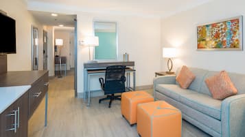 A hotel room with a desk, a sofa, an ottoman, a TV, and a painting on the wall.