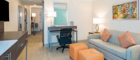 A hotel room with a desk, a sofa, an ottoman, a TV, and a painting on the wall.