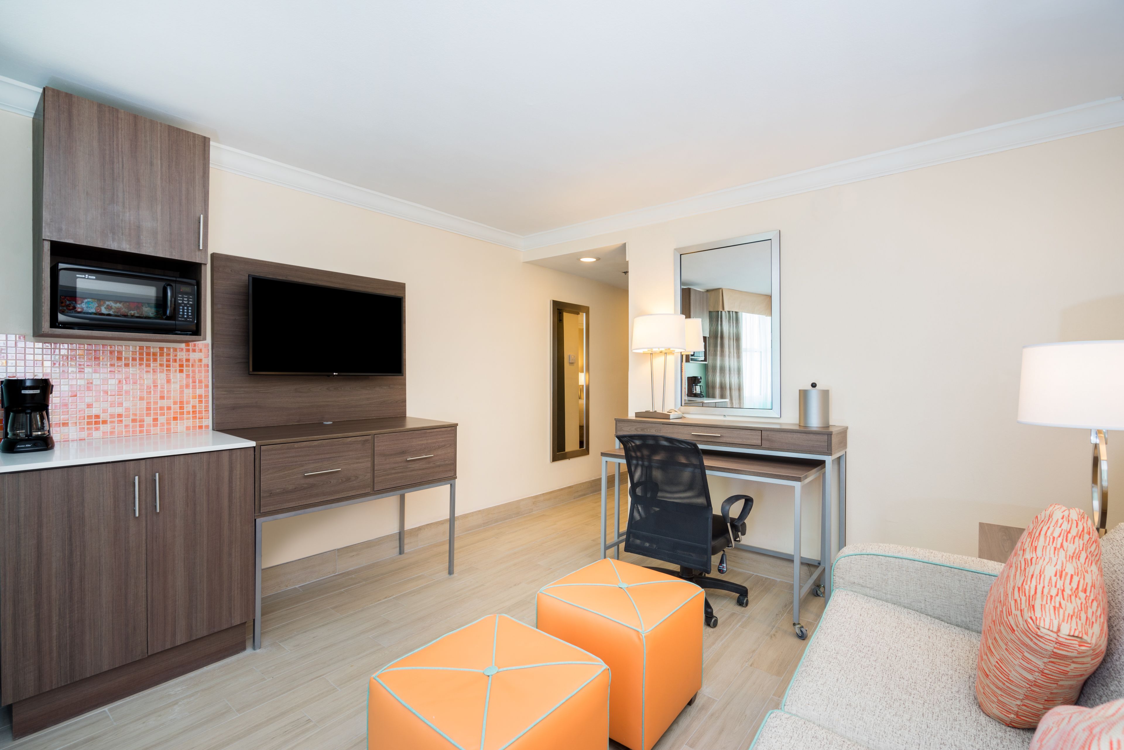 A modern hotel room with a kitchenette, a flat-screen TV, a desk with a chair, and a sofa with orange ottomans.