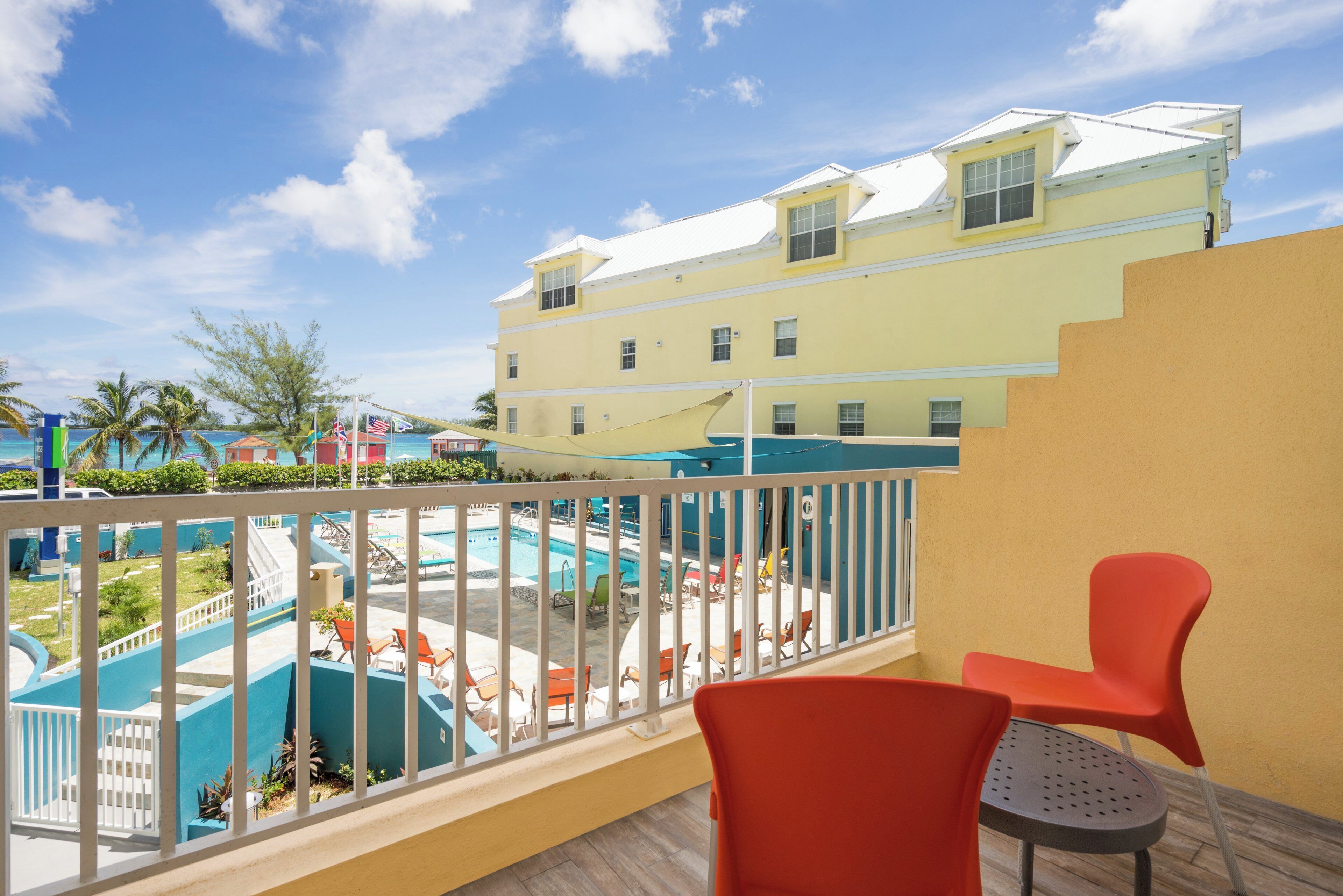 A balcony with a view of a swimming pool, a yellow building, and a clear sky.