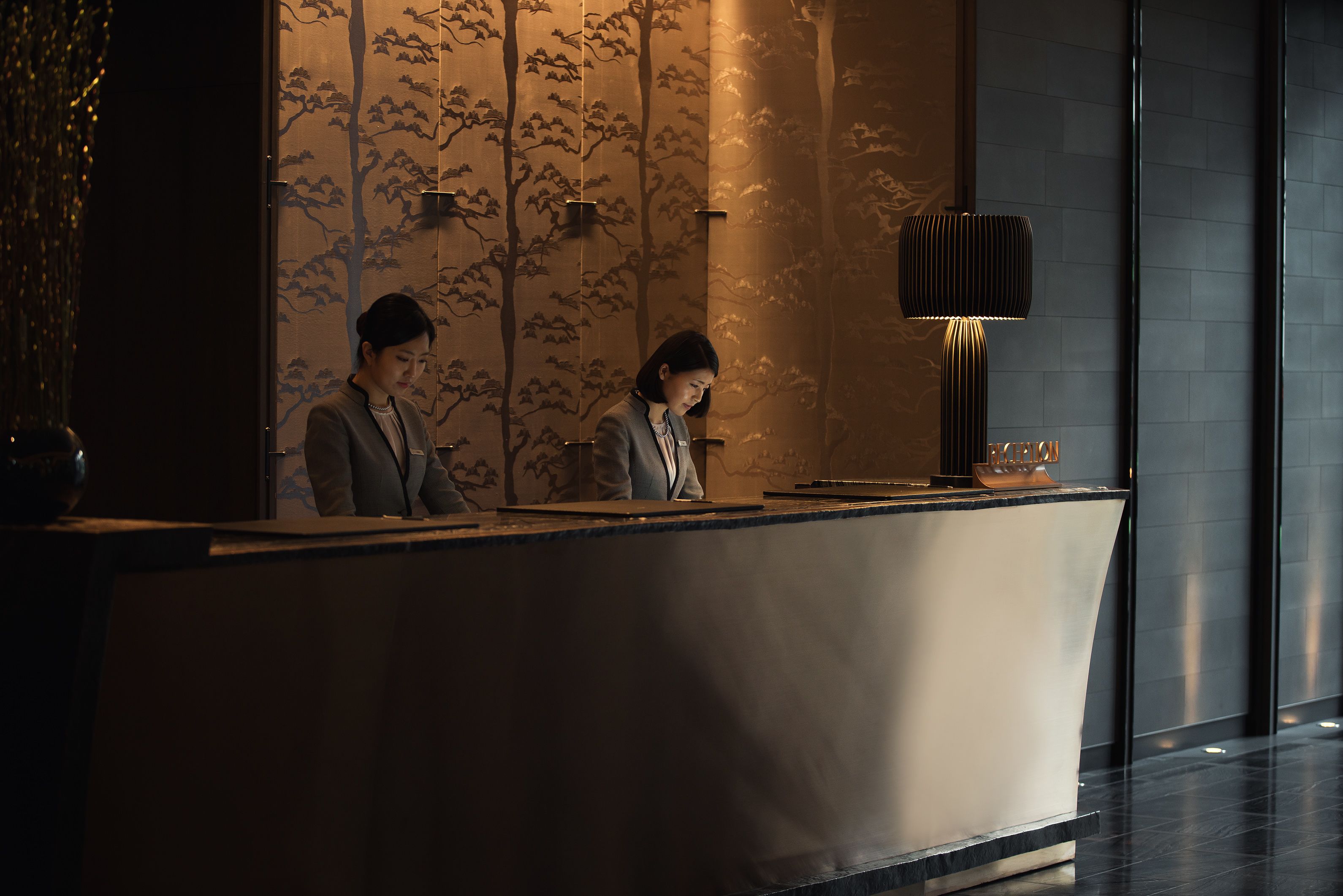 A reception desk with two staff members, a modern lamp, and a decorative wall with a tree pattern.
