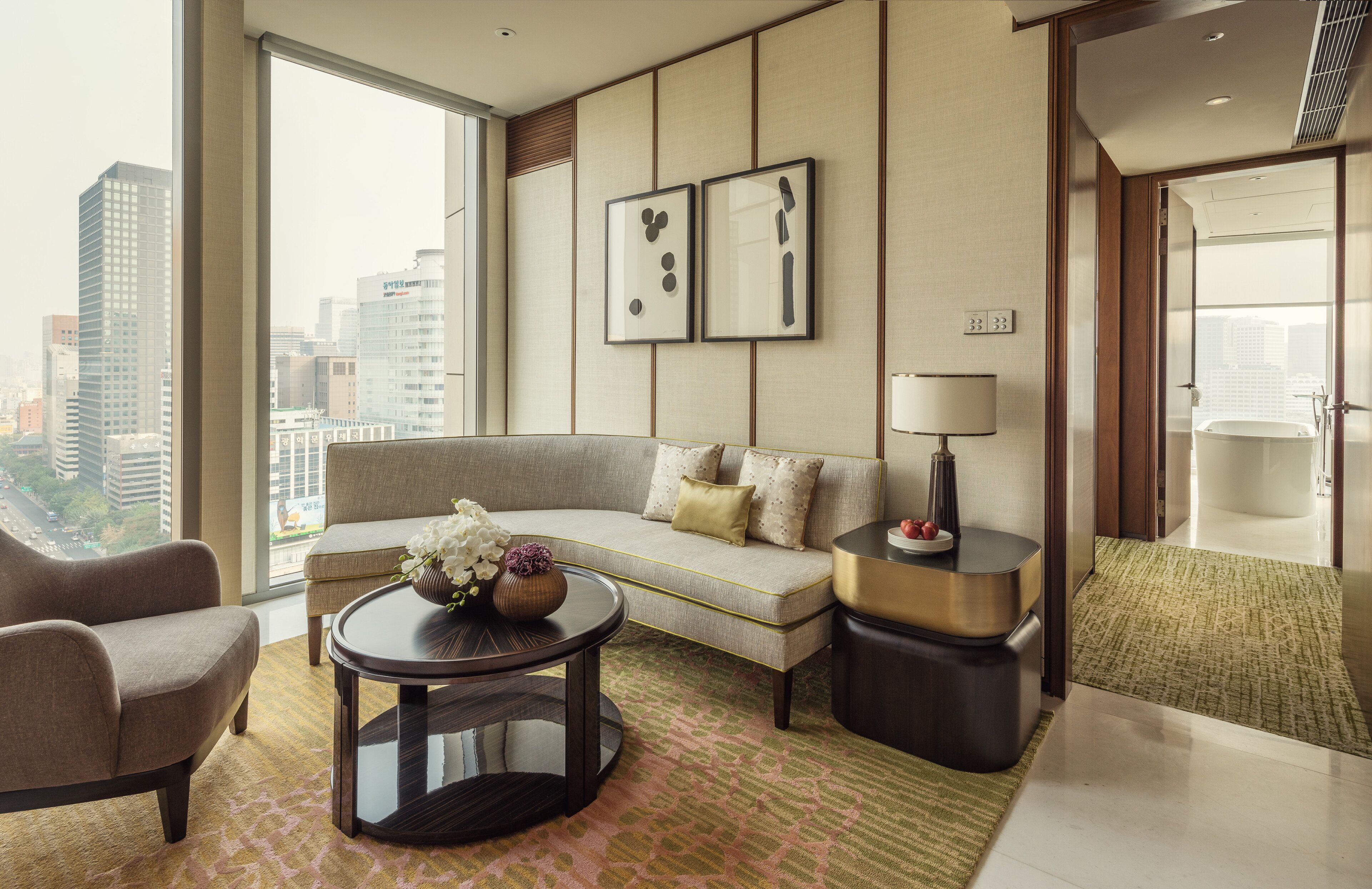 A modern hotel room with a sofa, armchair, and a round coffee table. There is a view of the city through large windows.
