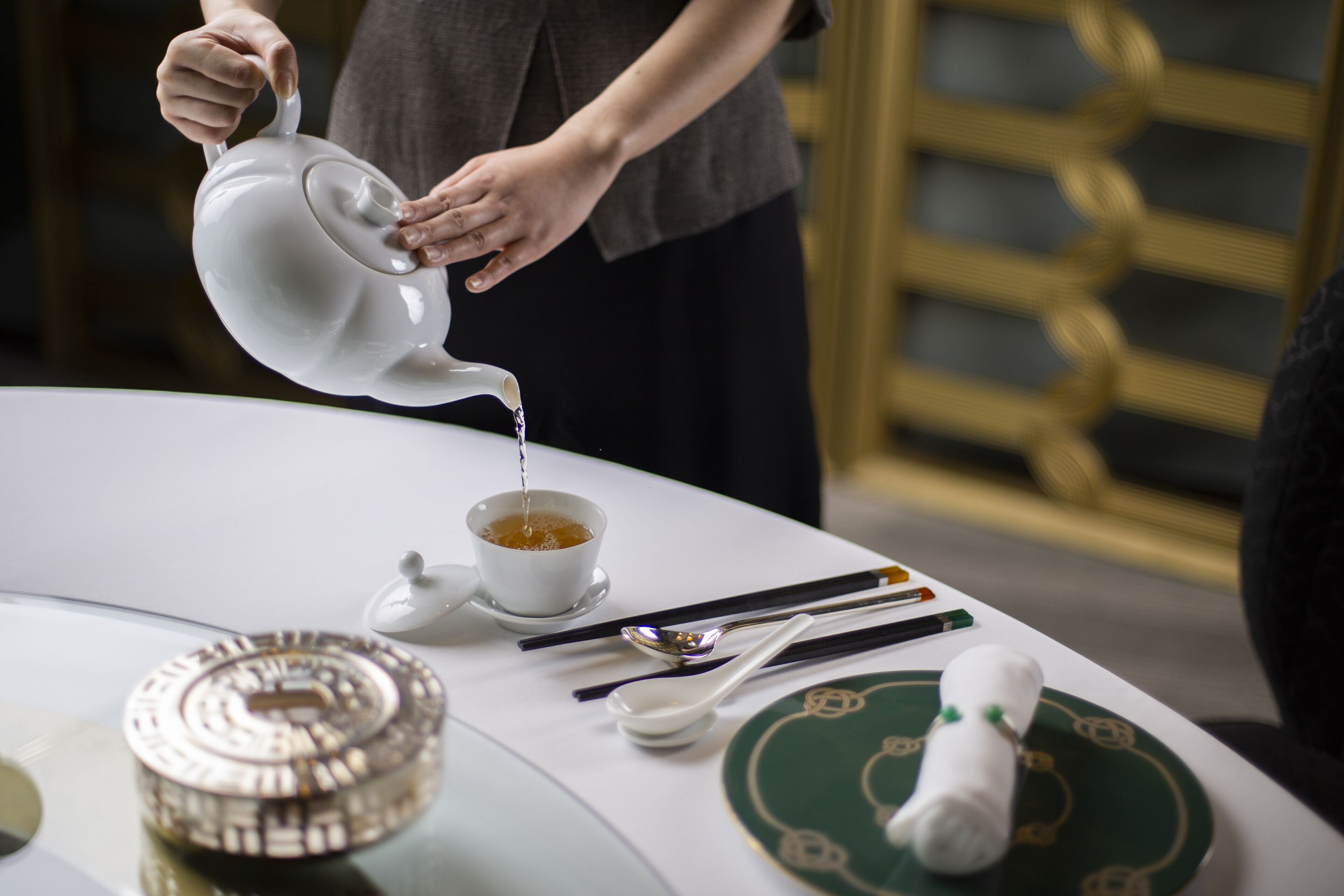 A person pouring tea into a cup on a table with chopsticks, a spoon, and a teapot.