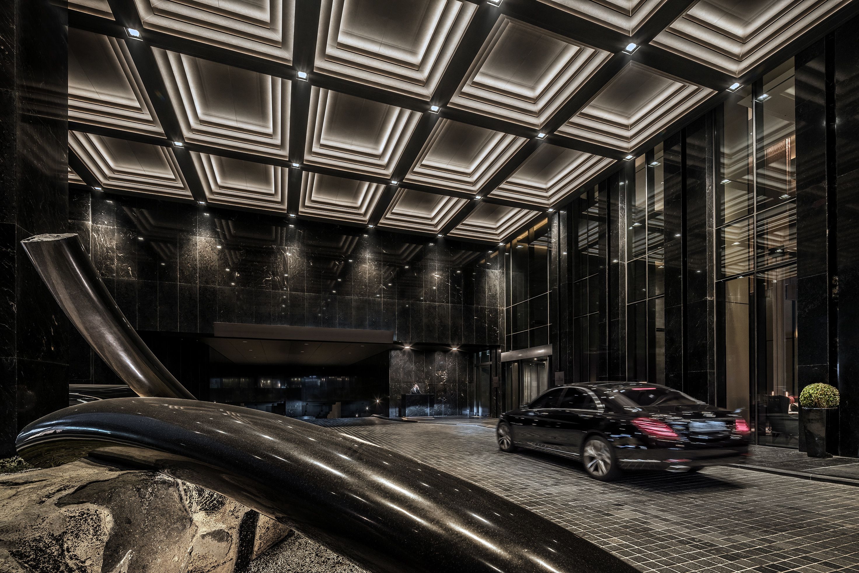 A modern building entrance with a black car parked outside, a glass facade, and a geometric ceiling design.