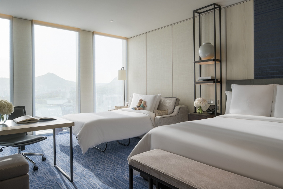 A hotel room with two beds, a desk with a chair, a lamp, and a nightstand with a vase of flowers.