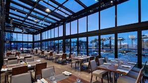 A restaurant with a glass ceiling, wooden tables, and chairs, and a view of palm trees and an airport in the background. - Hilton Garden Inn Izmir Bayrakli (Izmir)