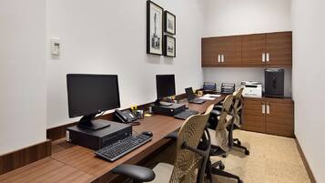 A conference room with a long wooden table, multiple monitors, laptops, and office chairs. There are framed pictures on the wall and a water dispenser on the right.